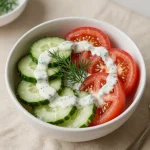 A vibrant creamy cucumber and tomato salad served in a bowl, garnished with fresh herbs.